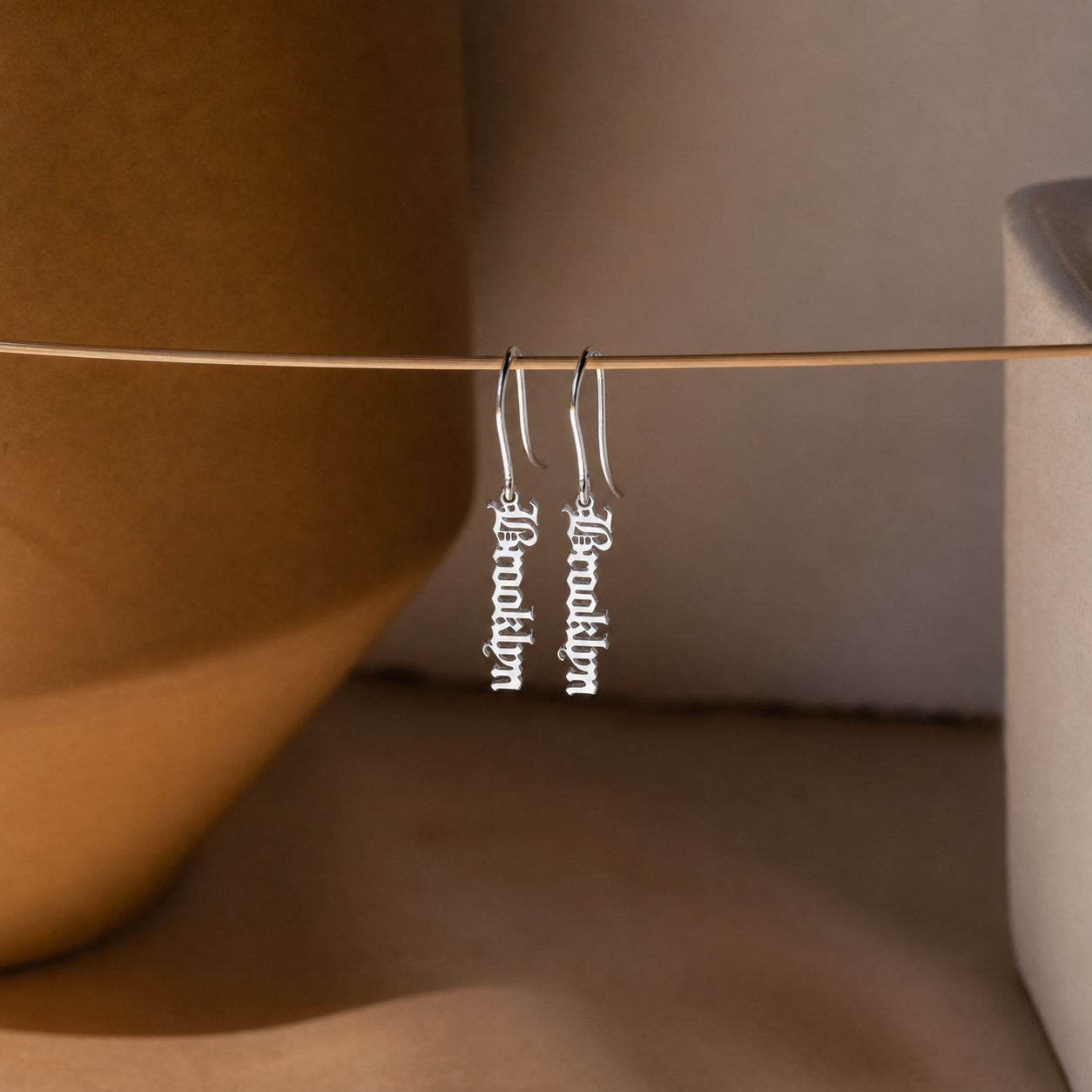Silver earrings with 'Brooklyn' inscription hanging on a stick against a neutral background
